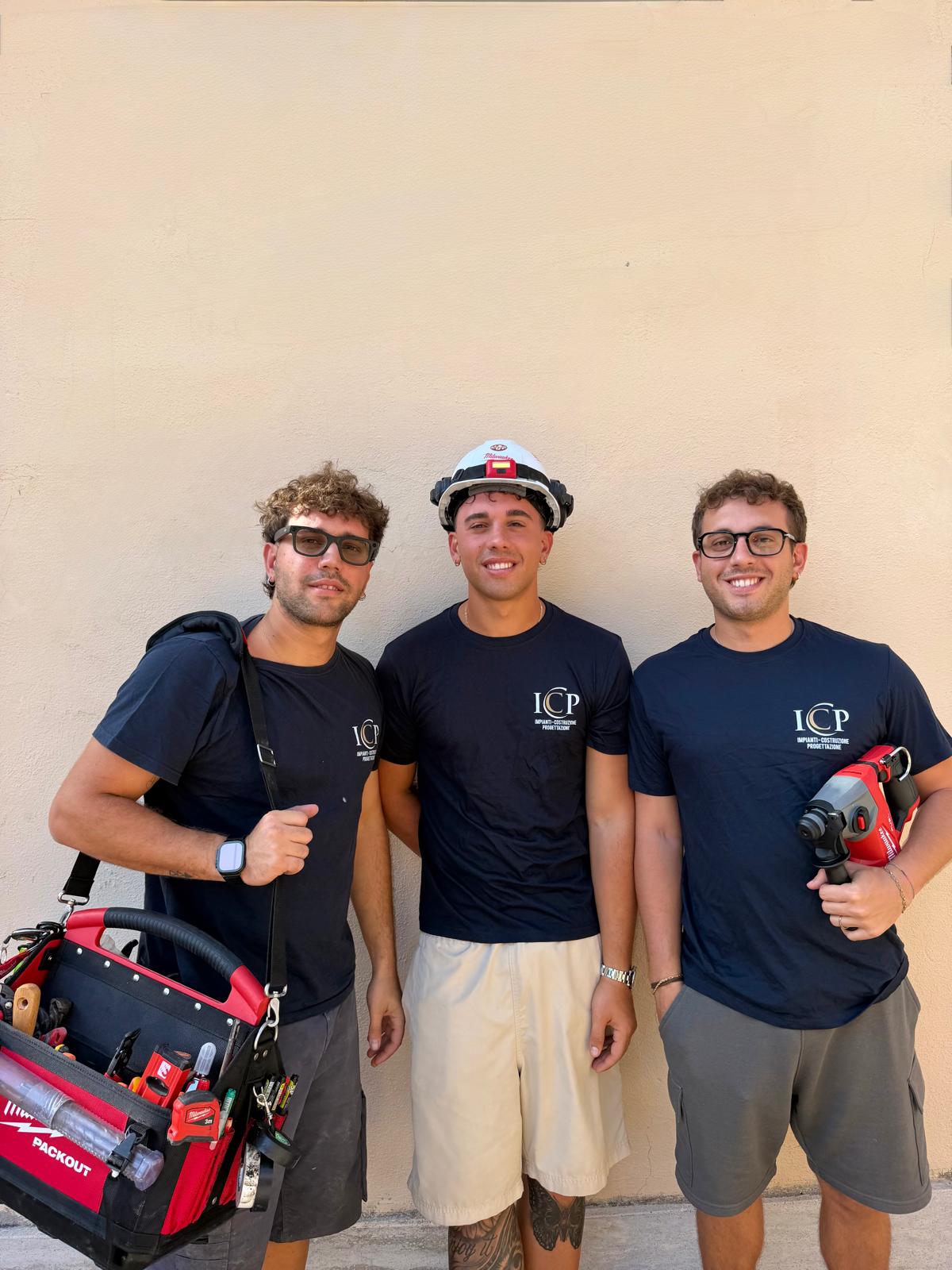 Tre ragazzi della squadra ICP con le magliette blu, il casco, la borsa attrezzi e un trapano